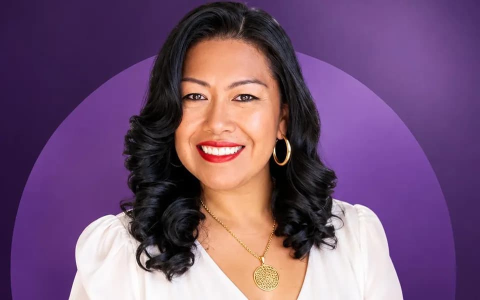 Lucy Peña sonriendo, con blusa blanca, sobre fondo morado